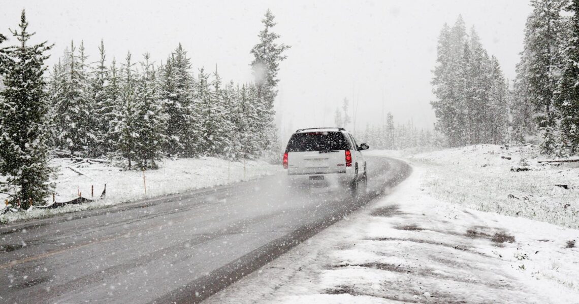 A car drives on a snowy road surrounded by trees during a snowstorm, creating a wintry scene.